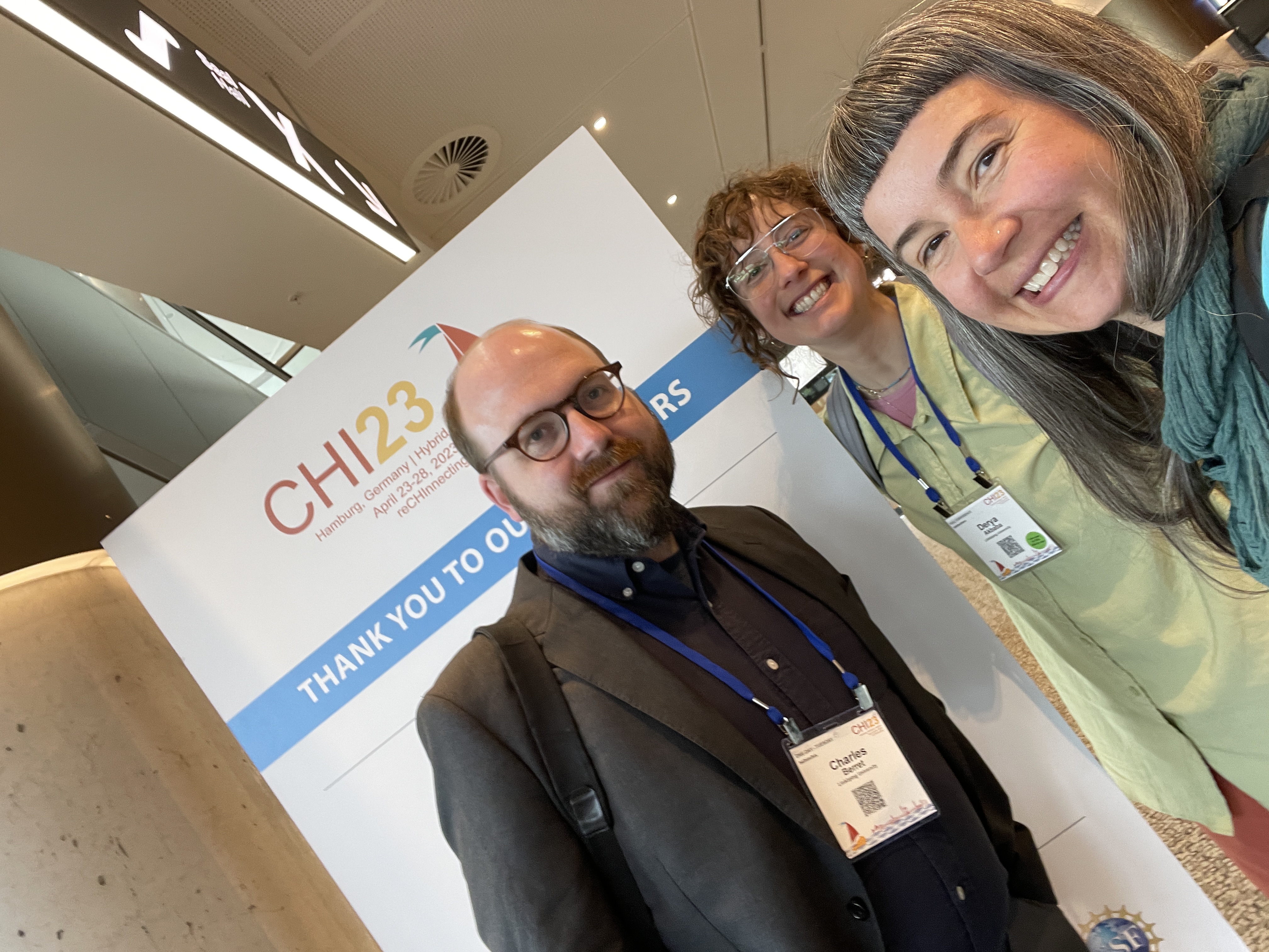 Charles, Derya, and Miriah smiling together in front of a CHI23 banner.